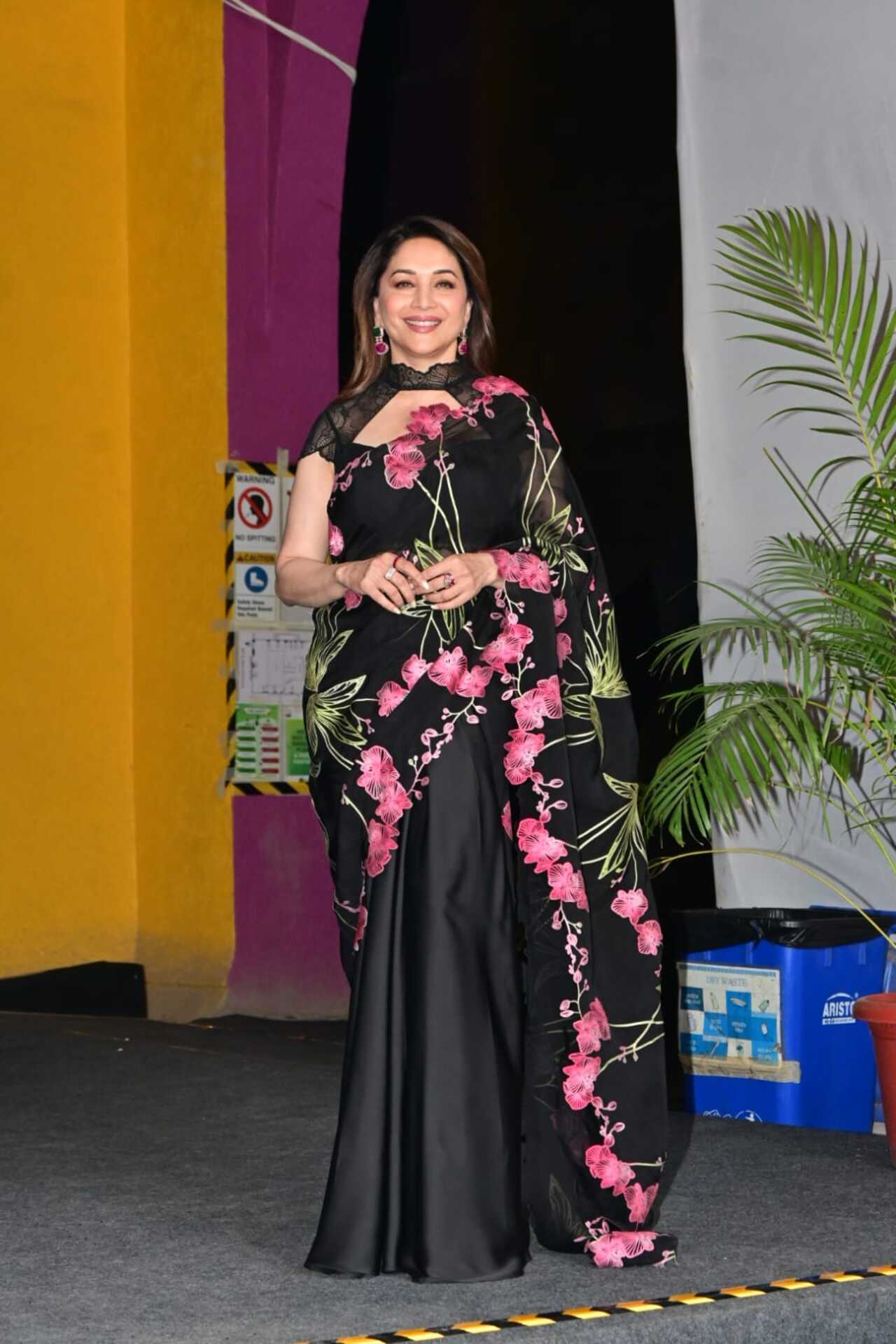 Madhuri Dixit looks breathtakingly elegant in a striking black saree that features a cascading trail of vibrant pink floral embroidery and delicate green leaf motifs.